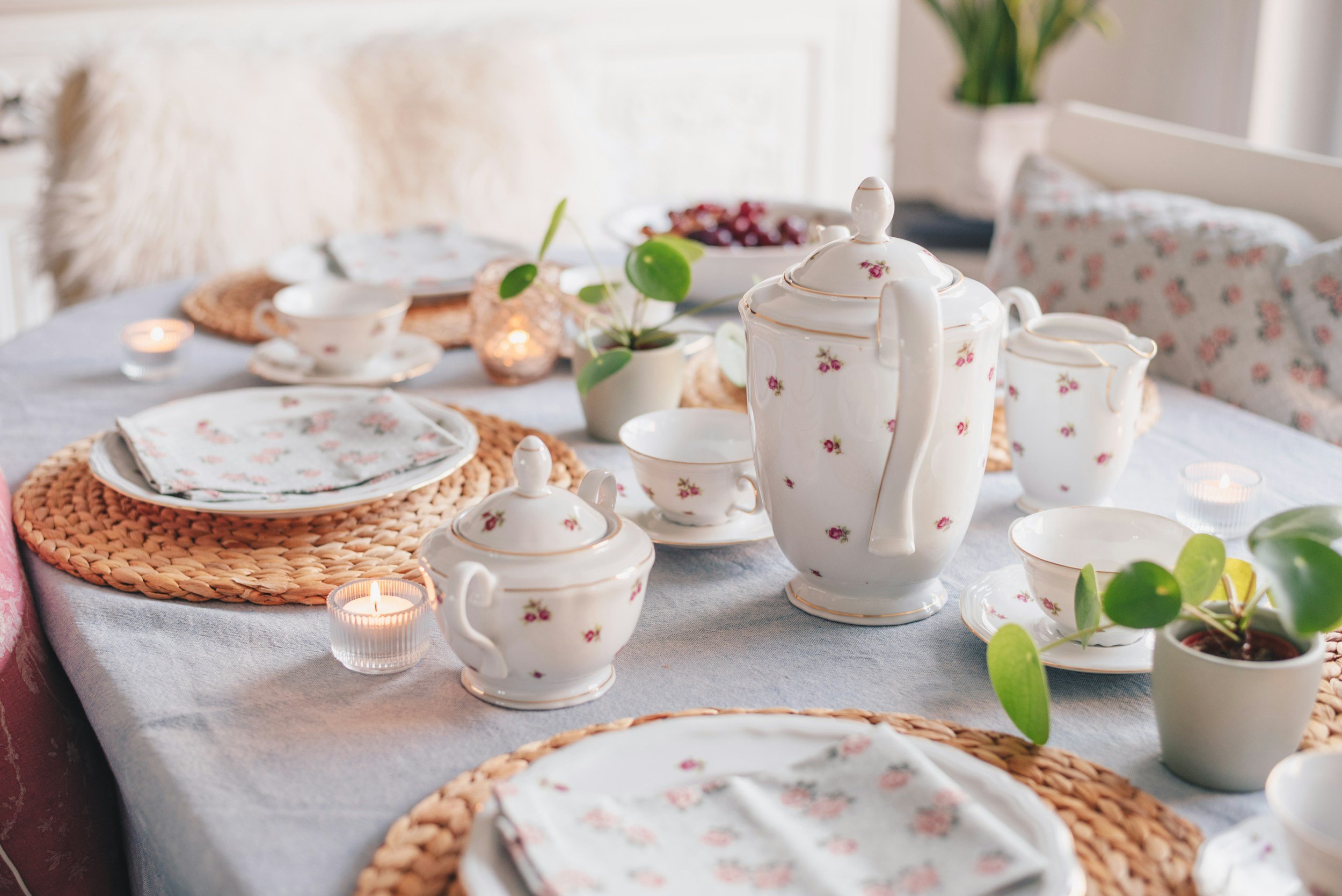 A tea party setup with floral patterned china, plates, teapots, cups, small candles, a plant, and a bowl of cherries on a table with a light-colored tablecloth in a cozy, sunlit room.