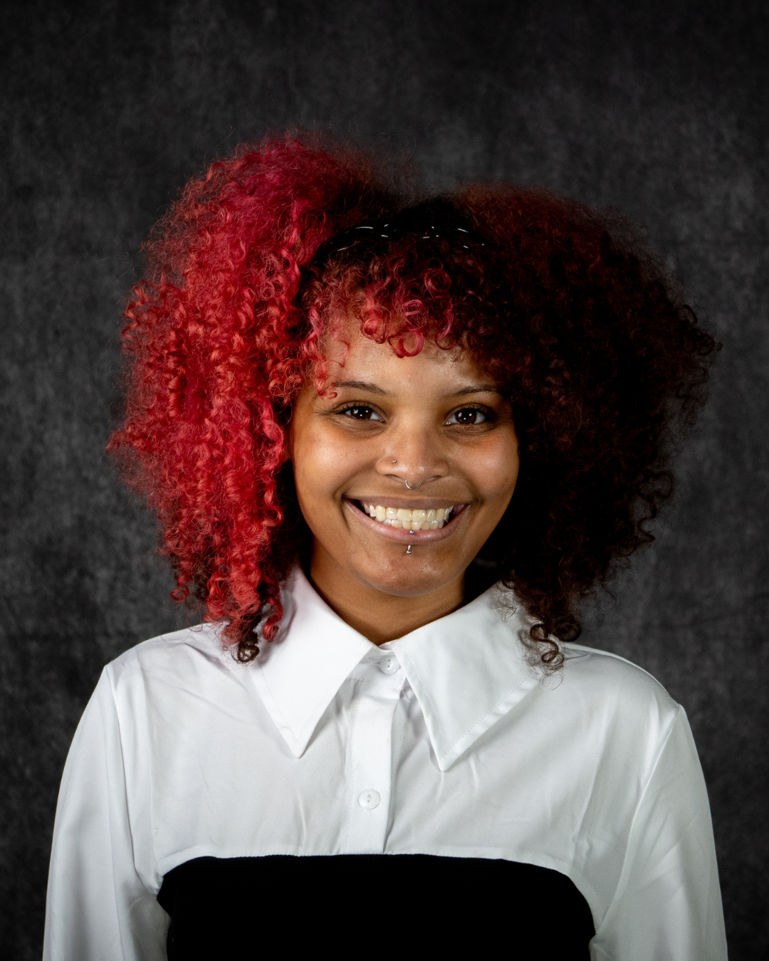 A person with curly hair dyed red and black, smiling, wearing a white and black shirt, against a dark backdrop.