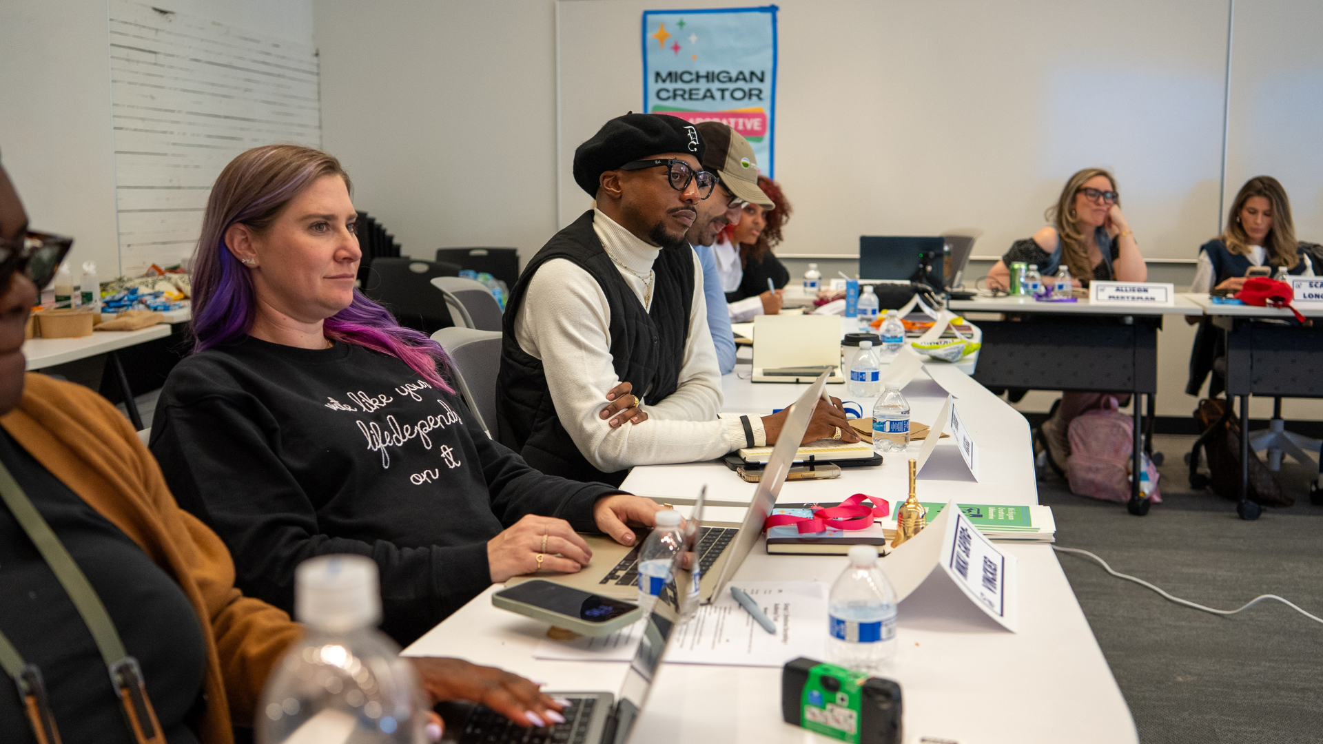 A group of people in a meeting room, sitting at a long table with laptops, notebooks, water bottles, and name cards, attending a presentation or workshop.