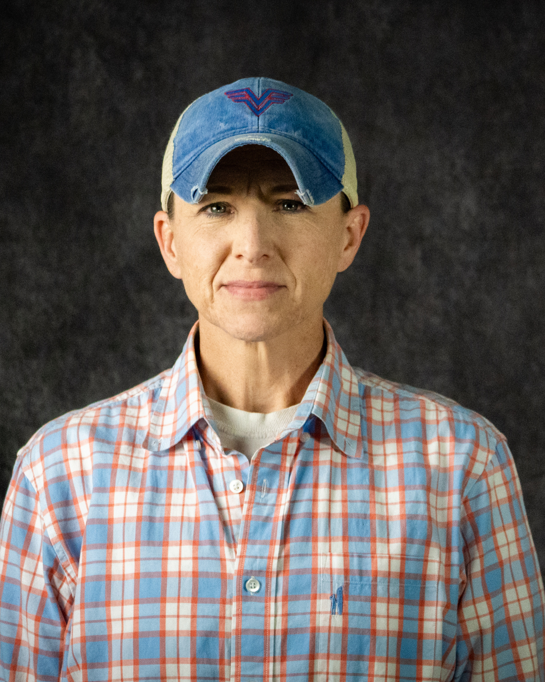 A person wearing a plaid shirt and a denim baseball cap with an embroidered logo, standing against a dark background.