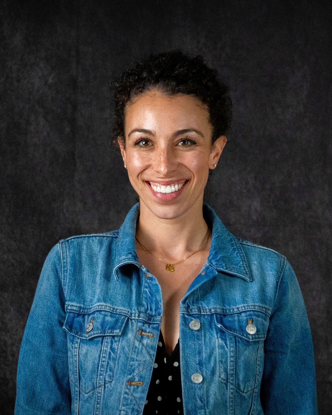 Smiling person with curly hair wearing a denim jacket and black polka dot top against a dark background.