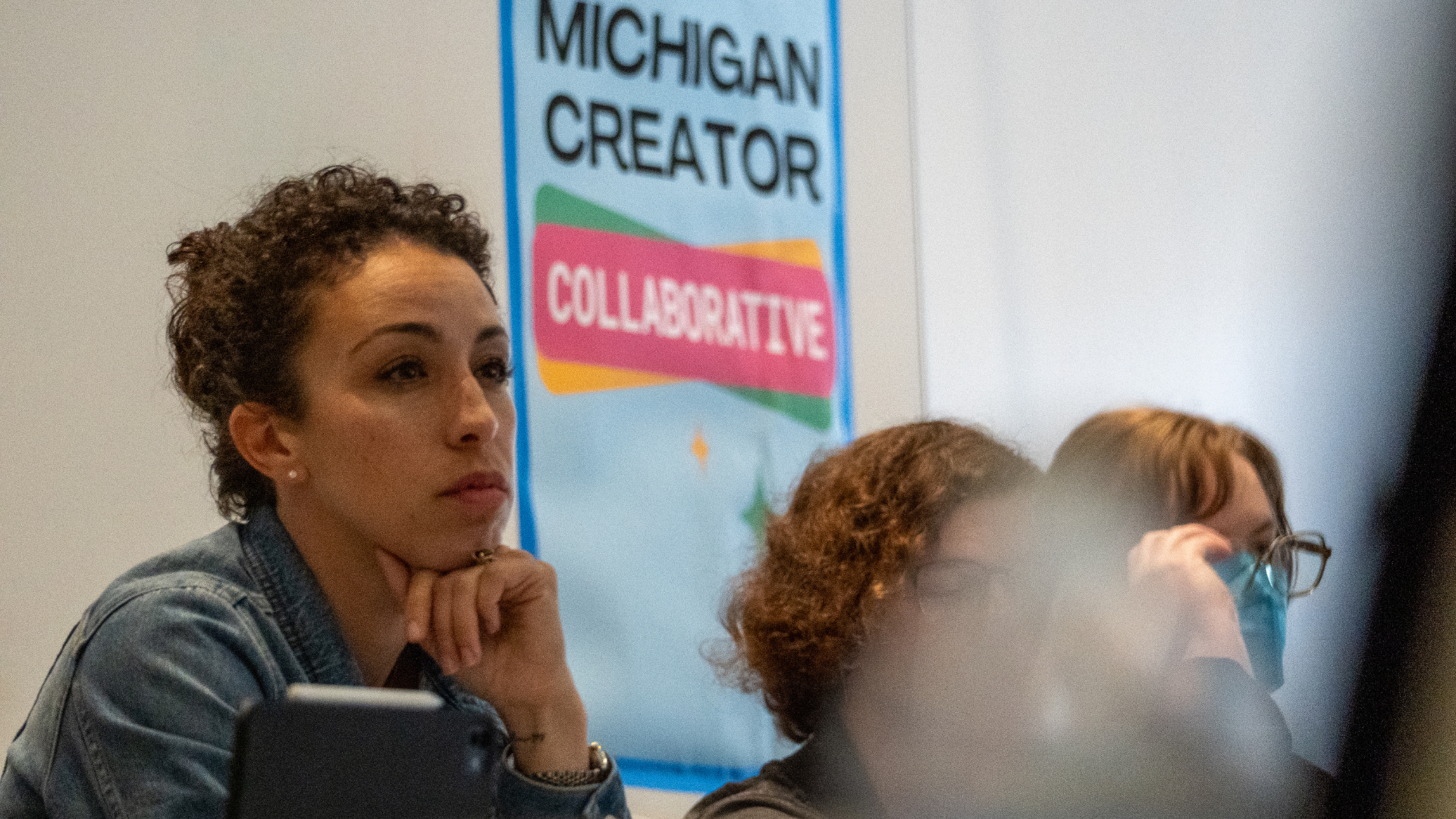 A woman with short curly hair, wearing a denim jacket, sitting with her hand on her chin. Behind her on the wall is a colorful sign that reads 'Michigan Creator Collaborative.' Other people are partly visible.
