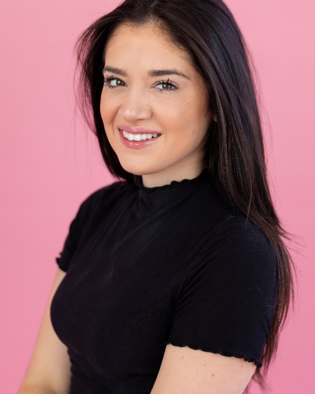 A person with dark brown hair smiling, wearing a black shirt, against a pink background.