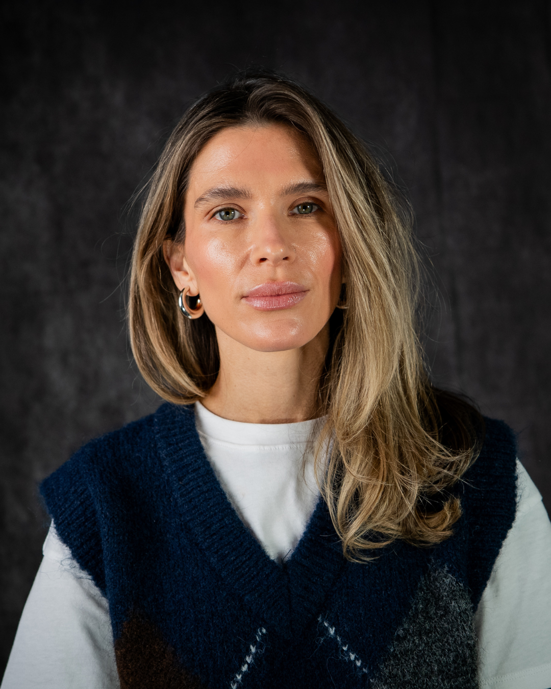 A person with shoulder-length blonde hair, wearing hoop earrings, a white shirt, and a navy blue sweater vest, poses against a dark background.