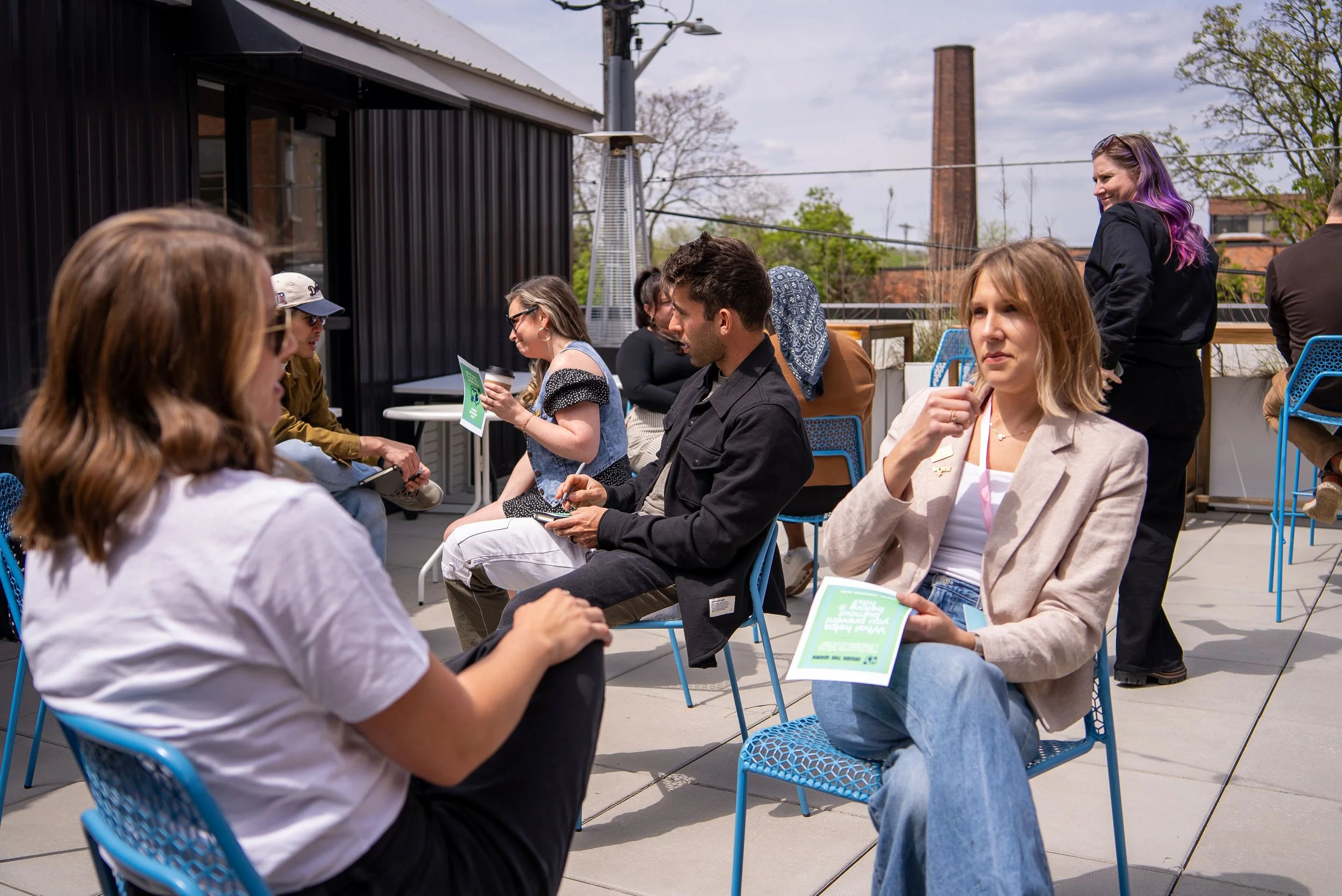 People sitting on chairs outdoors during a sunny day, engaged in conversations, with some holding papers or notebooks, and one person standing with purple hair in the background.