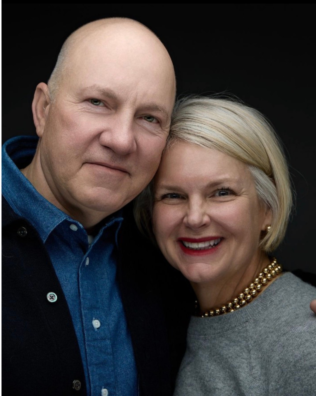 Two middle-aged people close together, smiling, with a dark background. Person on left has a bald head, light skin, and is wearing a blue shirt with a jacket. Person on the right has light skin, short blonde hair, red lipstick, and a gold necklace.