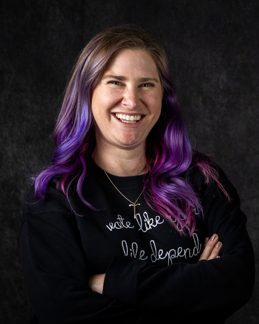 A person with purple wavy hair, wearing a black sweatshirt with white embroidered text and a gold necklace, standing against a dark background.