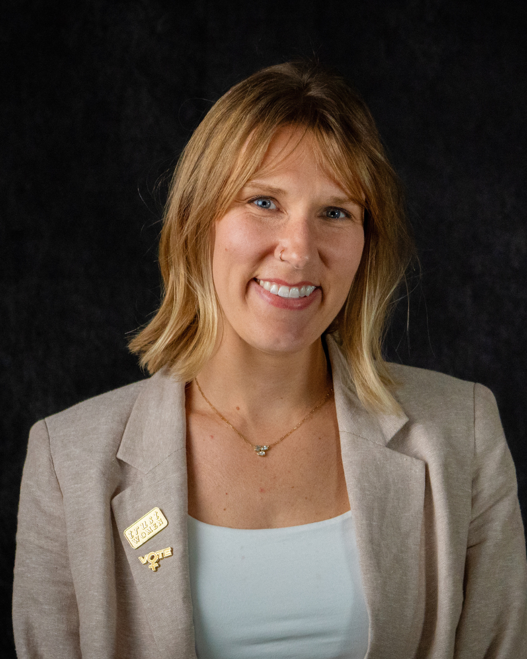 A person with shoulder-length blonde hair wears a beige blazer with a "Woman for Woman" pin and a white top. They have a necklace and a nose ring, and is smiling against a black background.