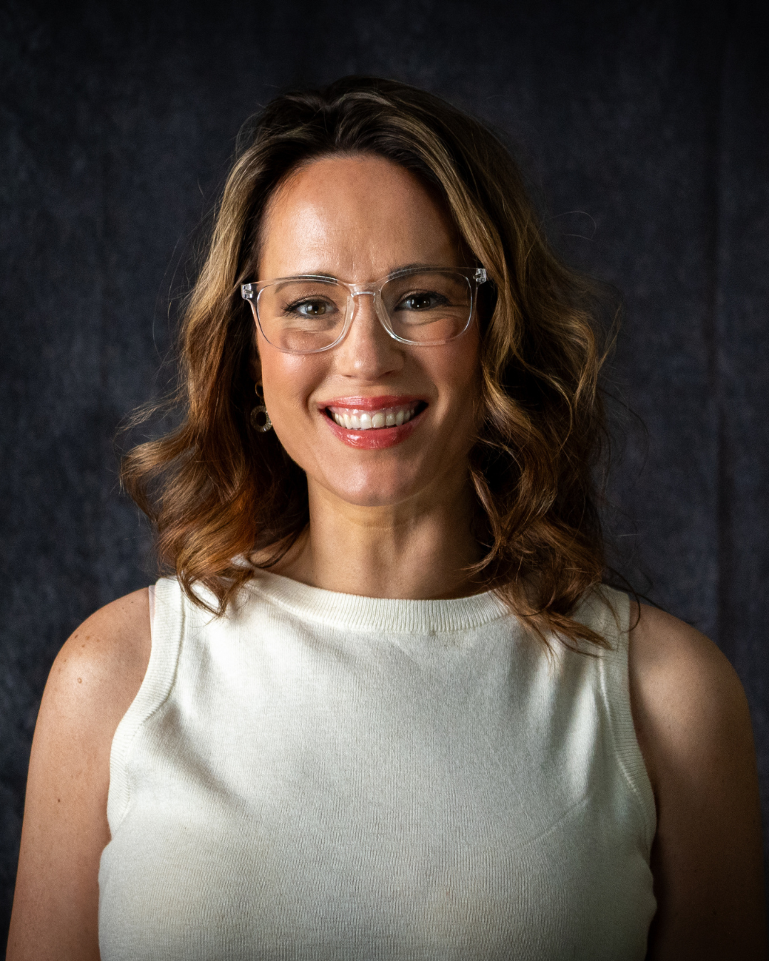 A person with wavy brown hair, wearing glasses and a white sleeveless top, smiling at the camera against a dark background.