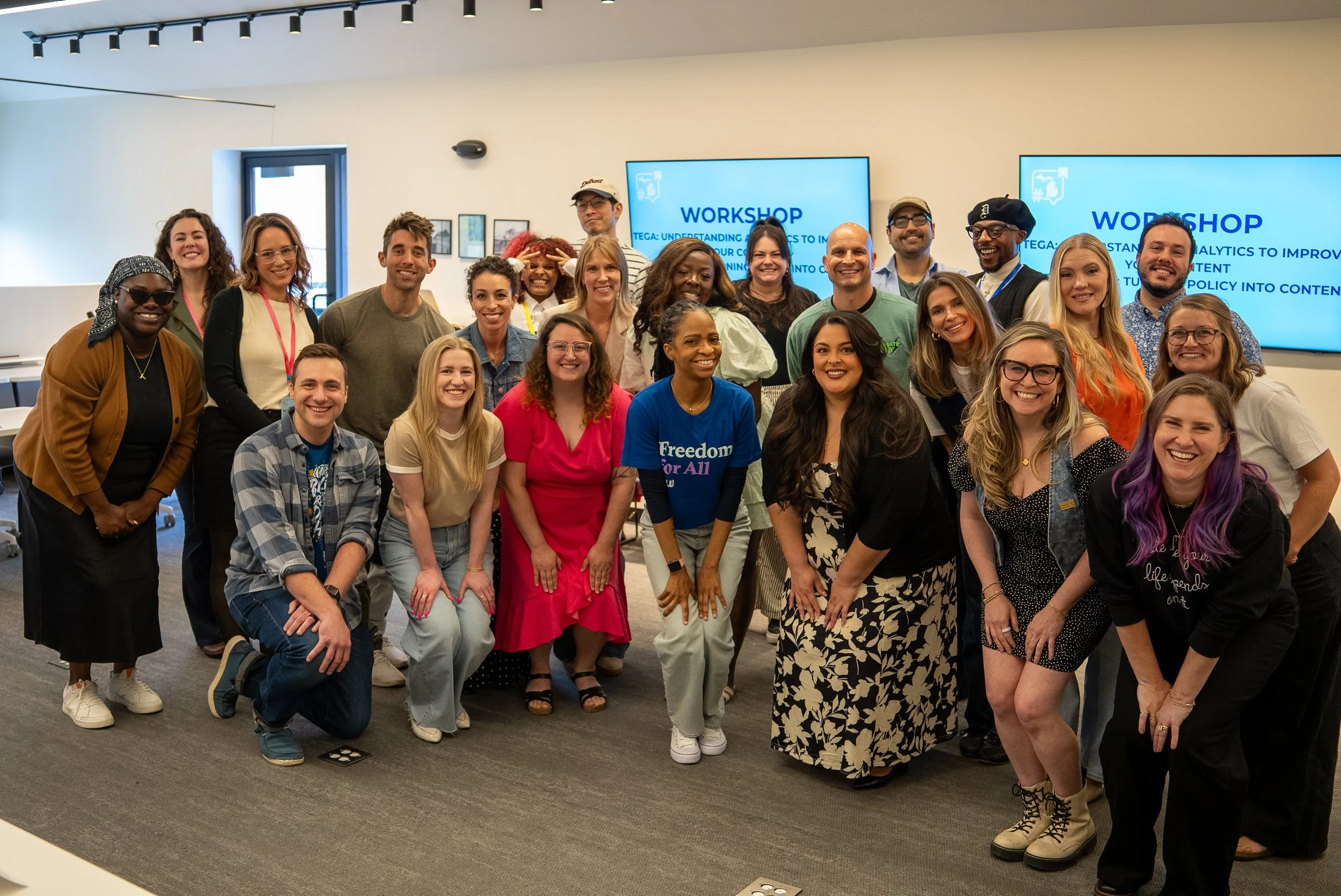 Group of diverse people smiling and posing for a photo at a workshop event, with screens displaying workshop information in the background.