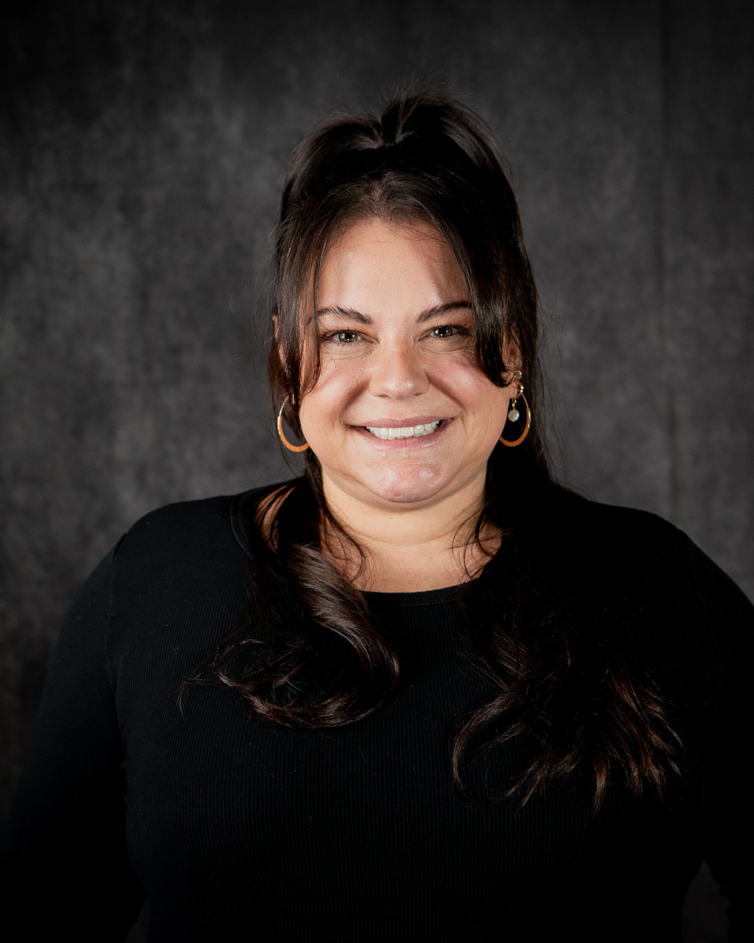 A person with dark hair and light skin, smiling, wearing hoop earrings and a black top, pose against a dark background.