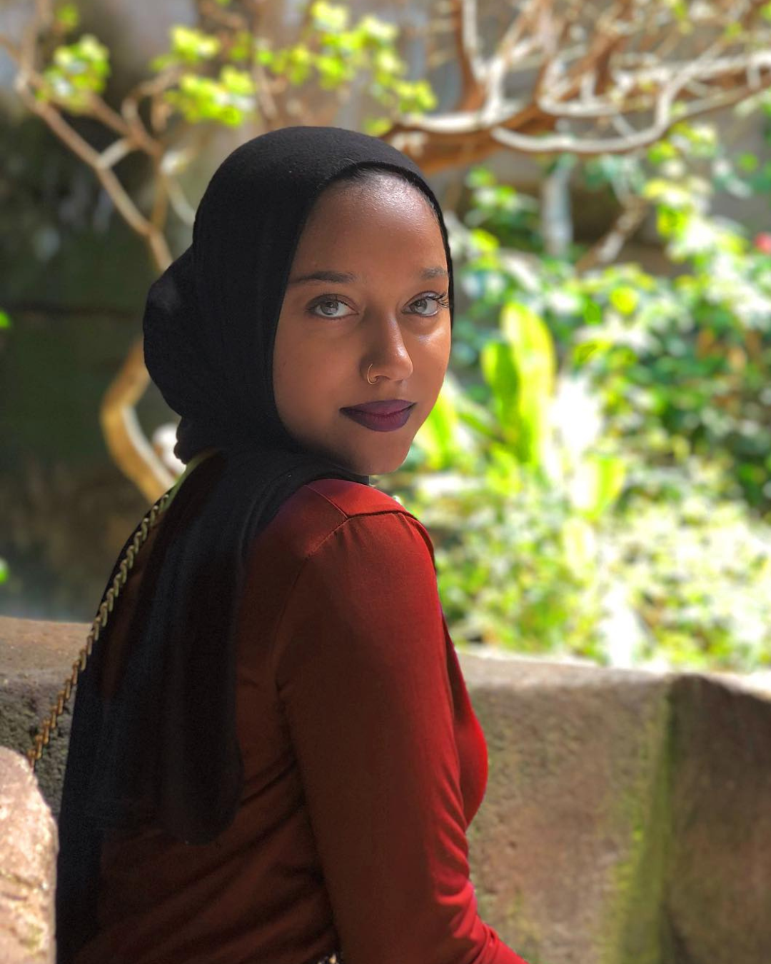 A person wearing a black headscarf and a red top, sitting outdoors on a stone surface surrounded by green foliage.