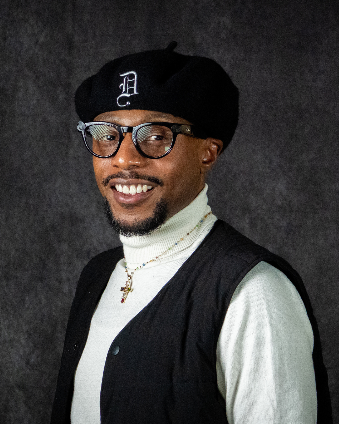 A smiling person wearing glasses, a black beret with a white letter D, a white turtleneck, a black vest, and a necklace, standing against a dark background.