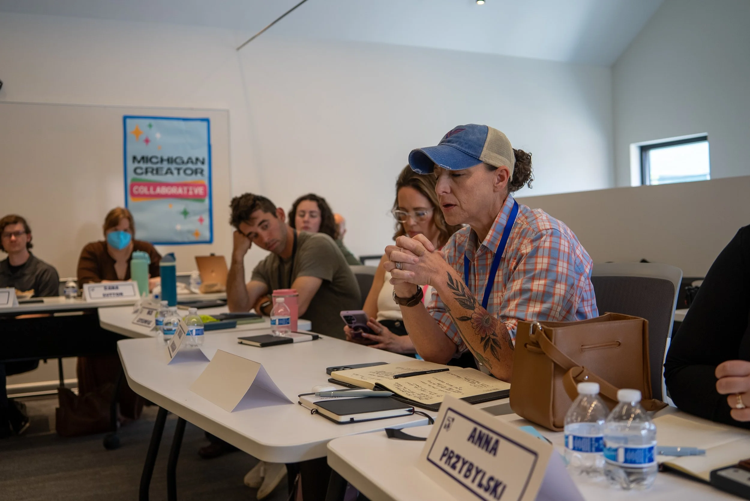 People sitting at a conference table, some with notebooks and phones, in a room with a sign reading 'Michigan Creator Collaborative' on the wall.