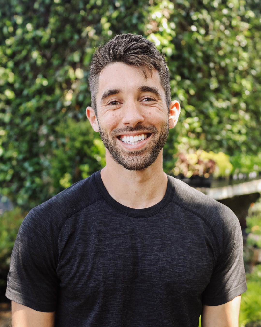 A smiling person with dark hair and a beard outdoors, wearing a black T-shirt, with green trees and plants in the background.