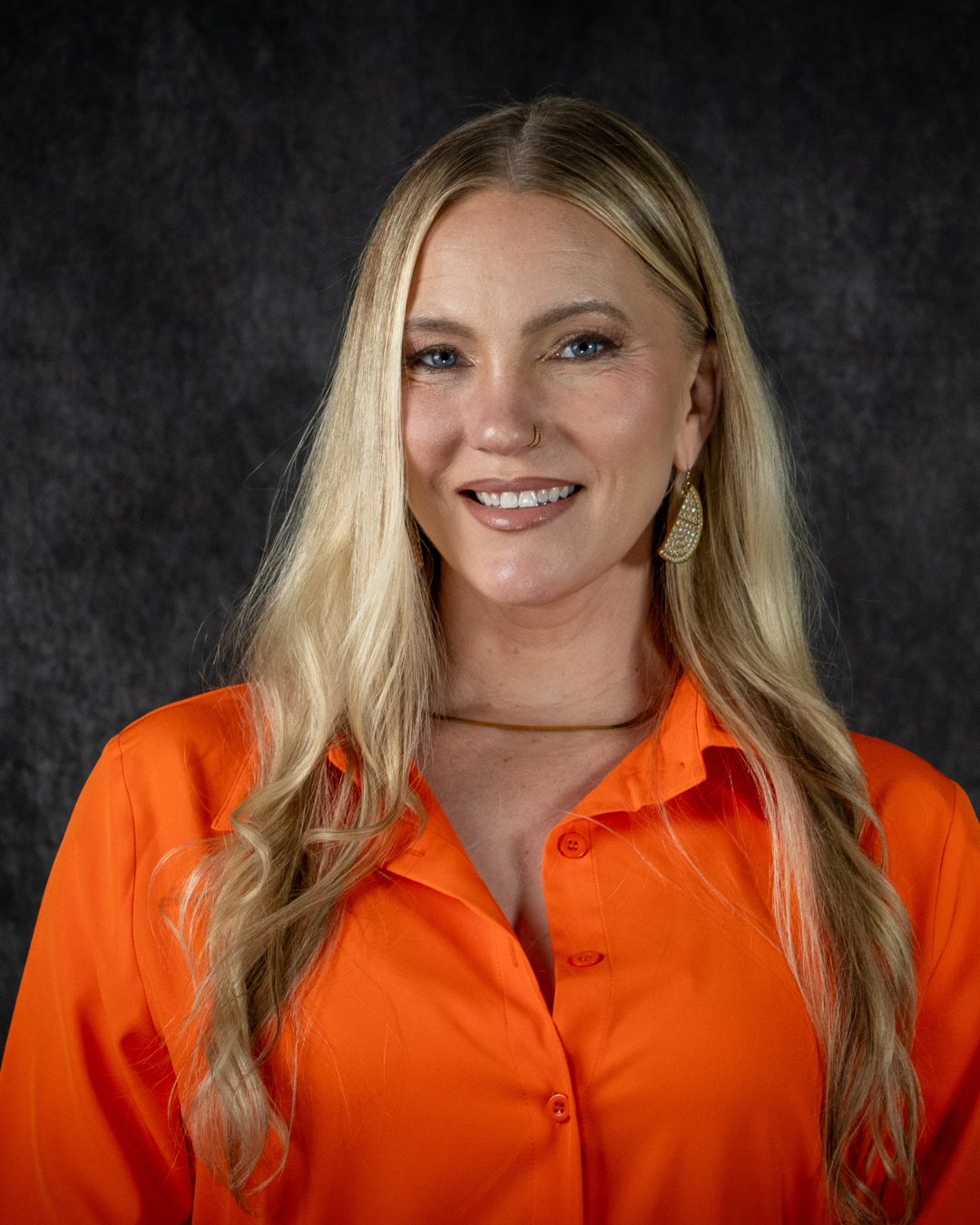 A person with long blonde hair wearing an orange shirt, gold jewelry, and a nose ring smiling against a dark background.