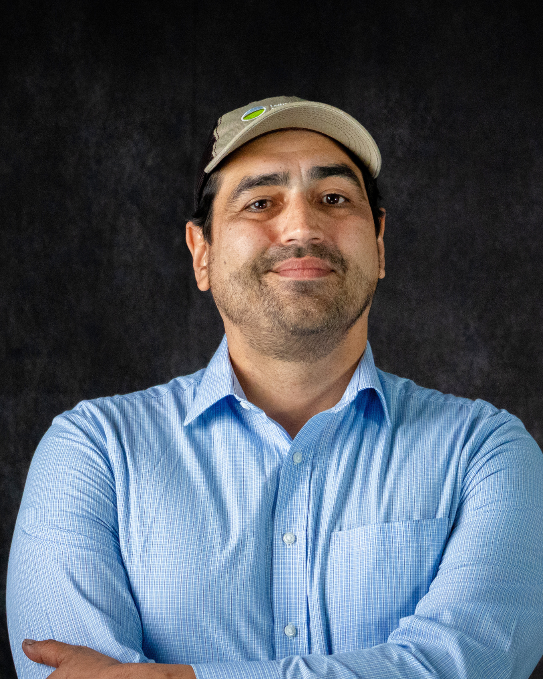 A person with a beard and mustache wearing a blue checkered shirt and a beige baseball cap smiling against a black background.