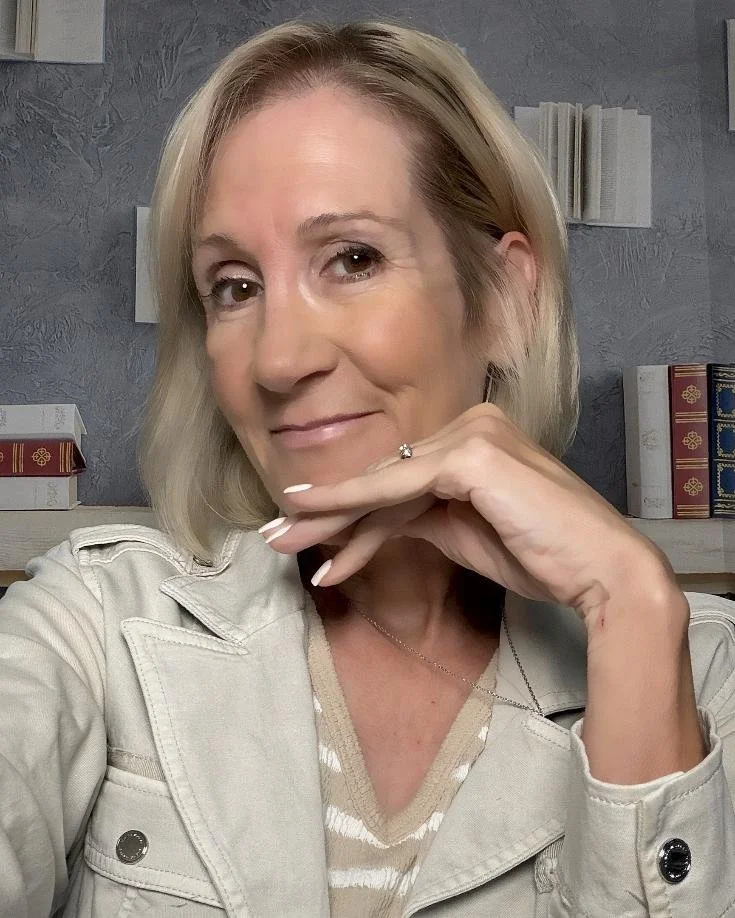 A person with blonde hair smiling, wearing a cream color, against a background with a few books and grey wall.