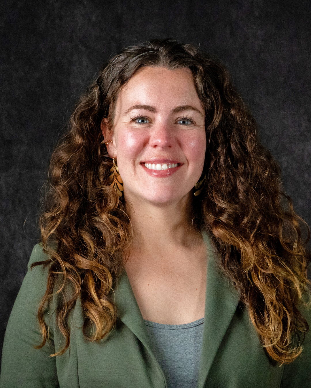 A person with long, curly brown hair and blue eyes, smiling, wearing gold earrings and a green blazer over a gray top, against a dark background.