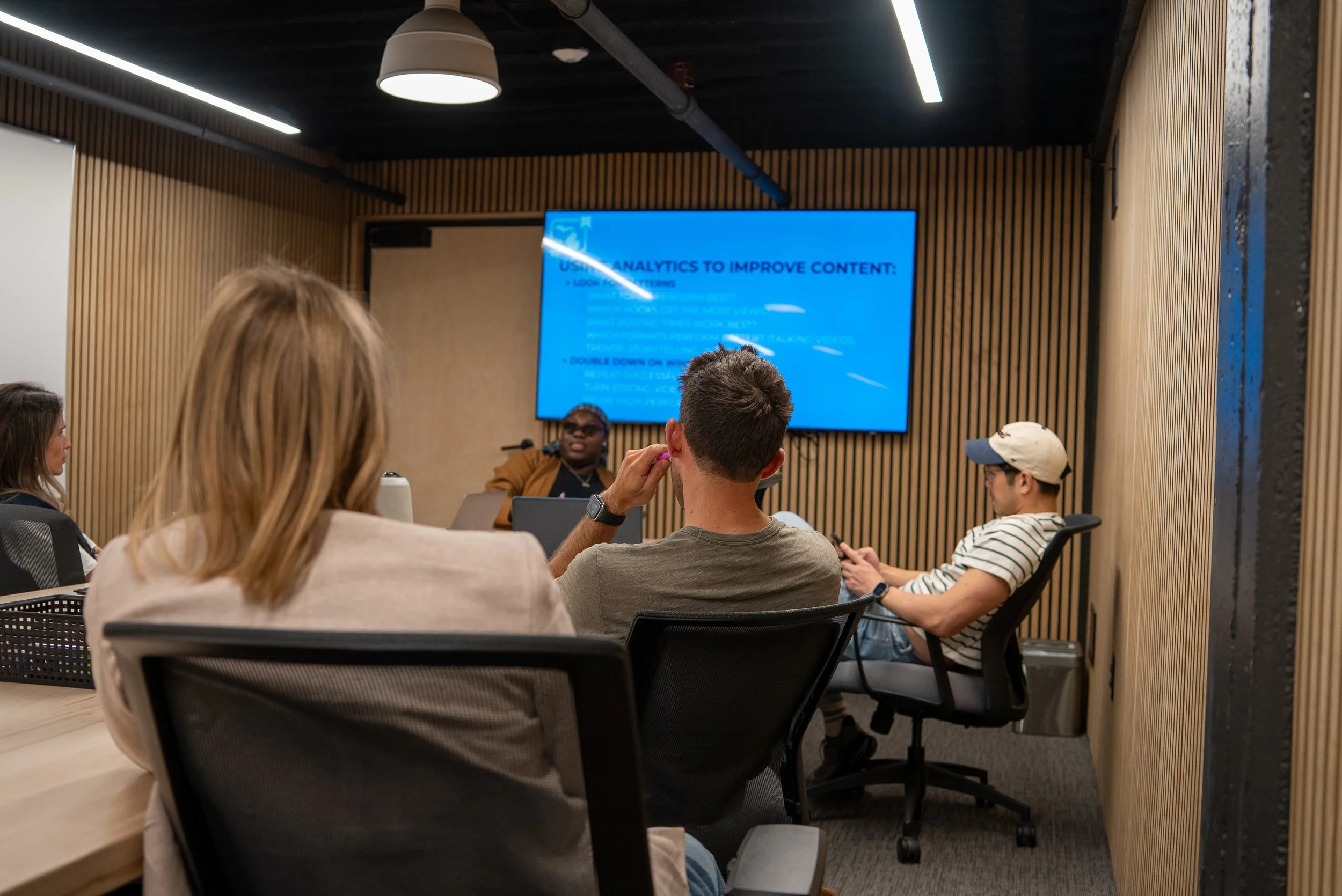 A group of diverse people in a conference room attending a presentation with a large blue screen displaying text about using analytics to improve content.
