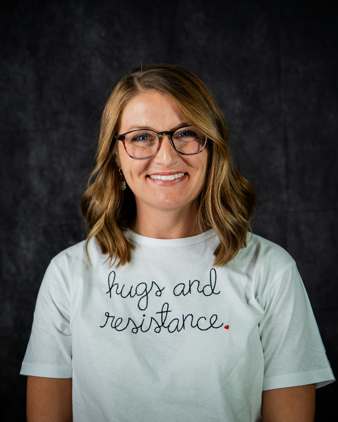 A person with shoulder-length brown hair, glasses, and earrings, smiling at the camera, wearing a white t-shirt with the message 'hugs and resistance' written on it.