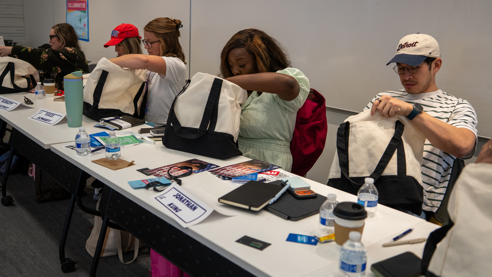 People sitting at a long meeting table with bags, water bottles, and pamphlets, reaching into bags, with name tags on the table.
