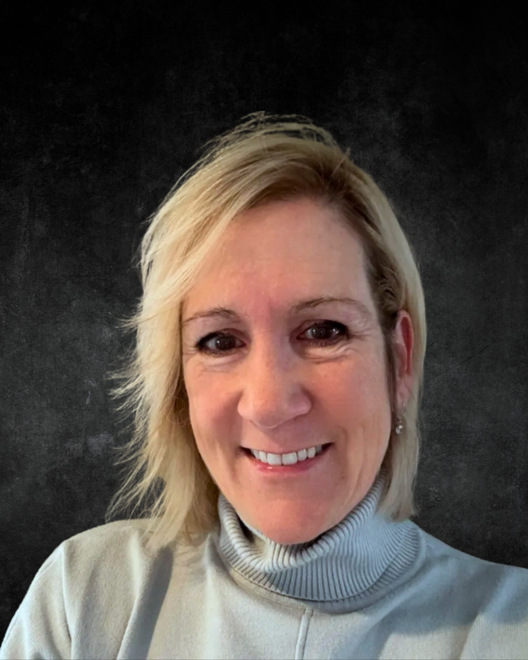 A person with blonde hair smiling, wearing a light grey turtleneck sweater, against a dark background.