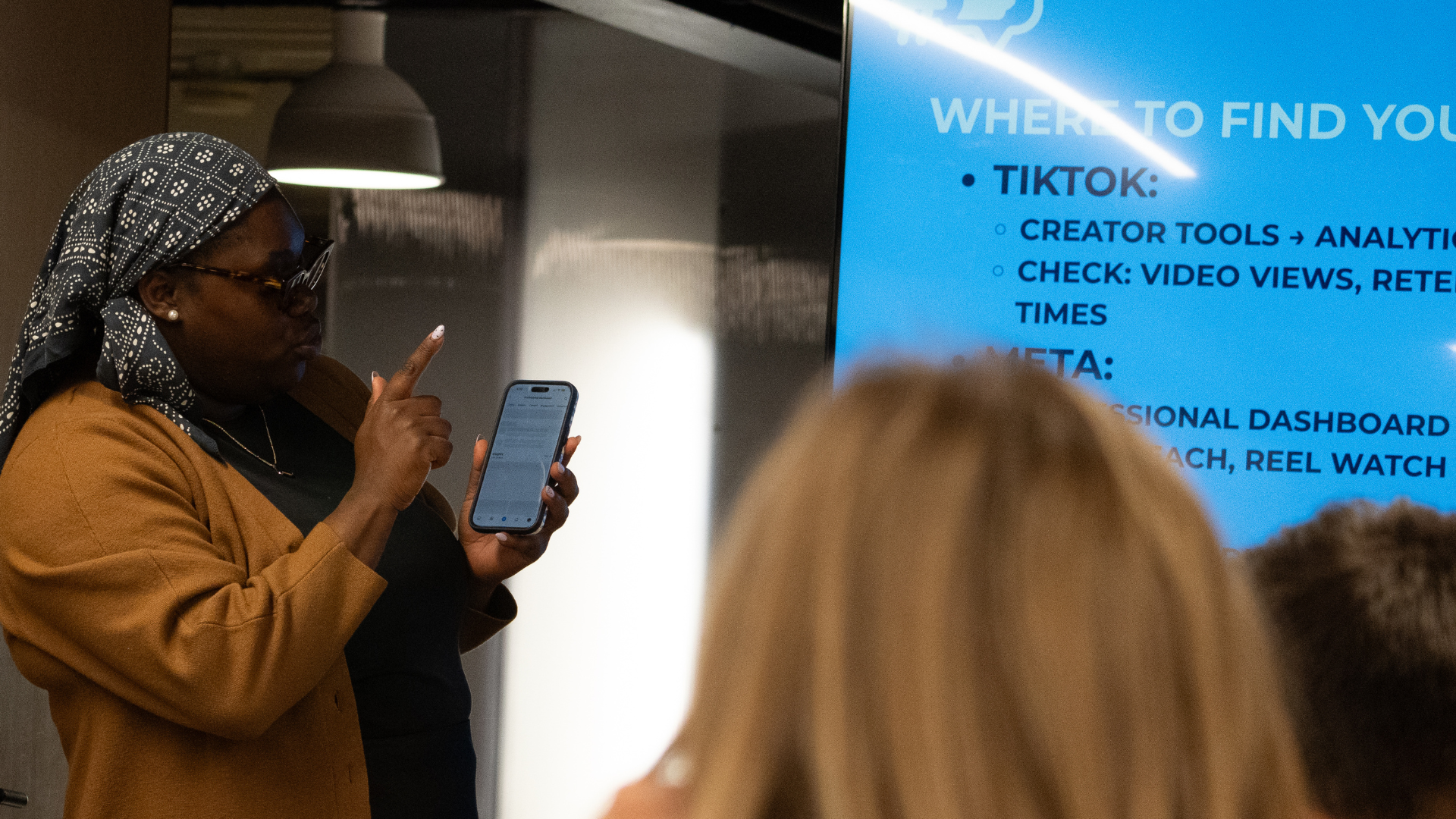 Woman in brown blazer wearing glasses and a patterned headscarf pointing upward while holding a smartphone during a presentation or meeting, with a digital screen displaying text about TikTok and analytics in the background.
