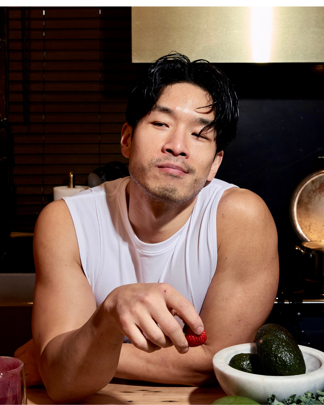 A person with black hair and slight facial hair wearing a white sleeveless shirt, sitting at a table holding a strawberry. There is a bowl of avocados in front of them and the background features wooden blinds and a lampholder.