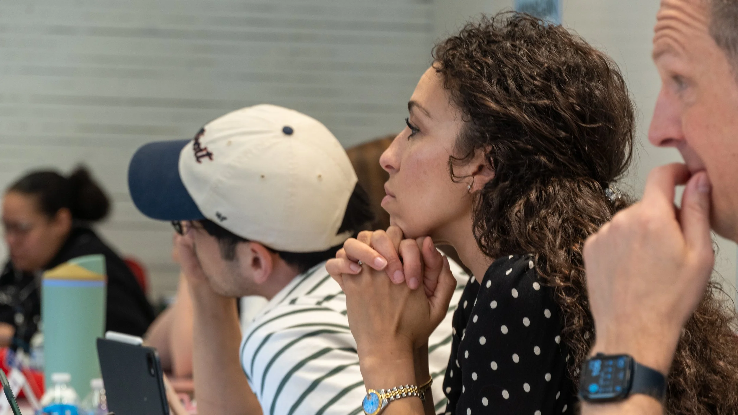 Group of people seated at a table, paying attention, with some resting their chins on their hands. One woman has curly hair and is wearing a black polka-dot shirt, while others are wearing striped shirts or hats. The setting appears to be a conference or meeting.