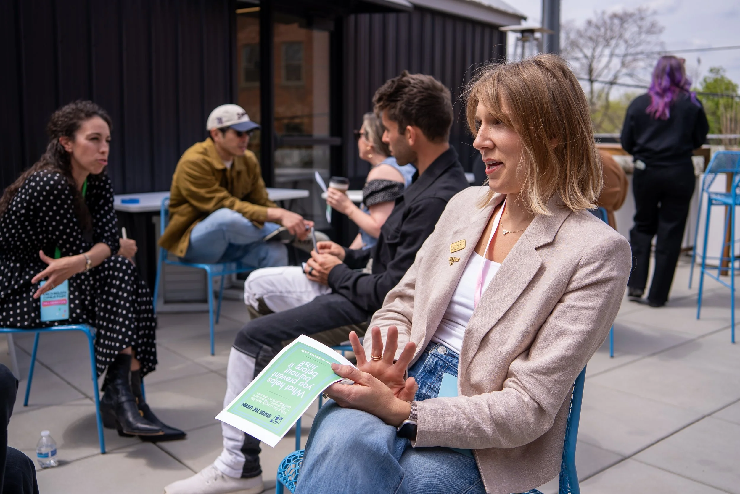 A woman with shoulder-length blonde hair wearing a beige blazer and jeans is sitting outside on a blue chair, holding a flyer about youth development. Other people are seated nearby and conversing at an outdoor event on a patio with black siding buildings in the background.