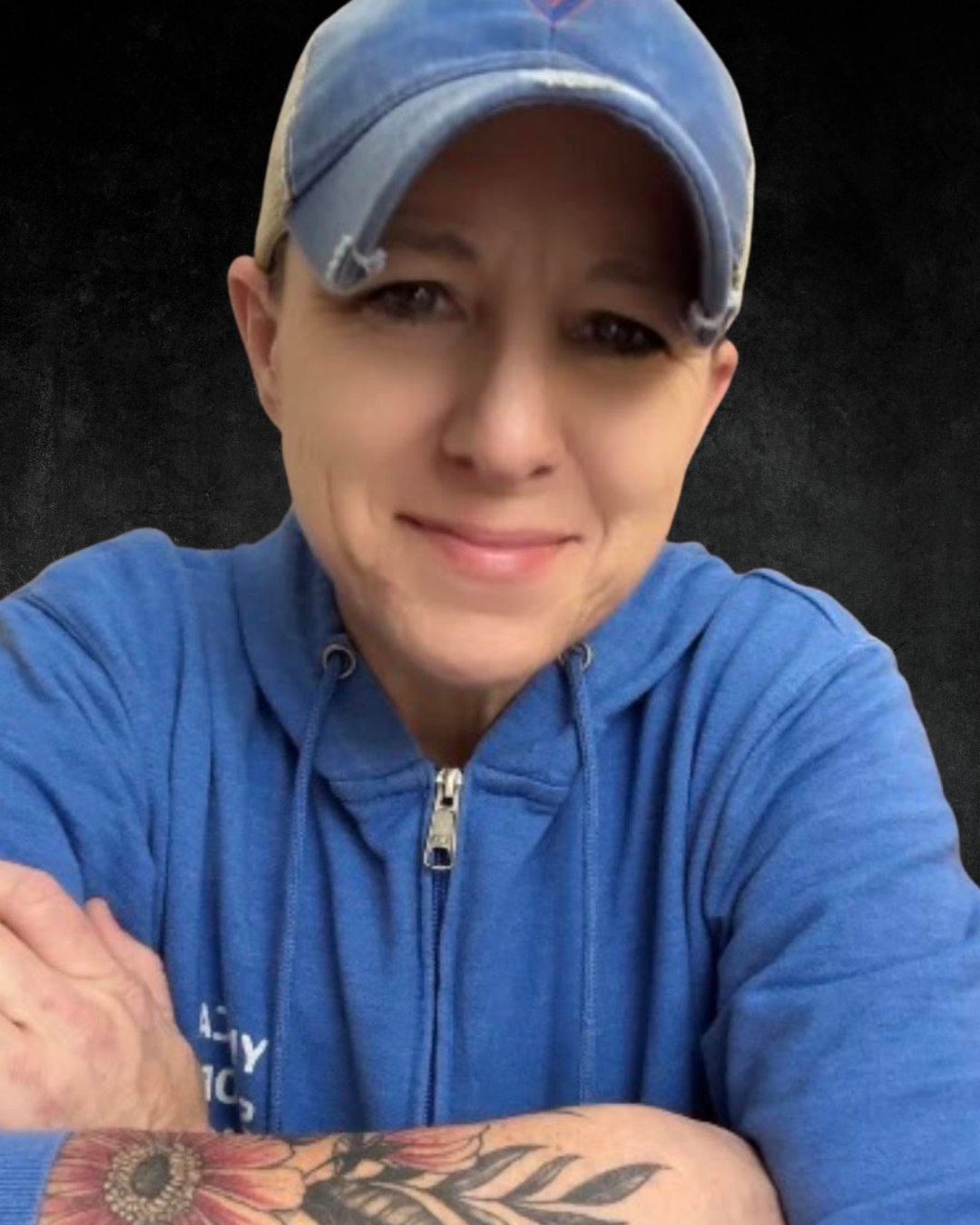 A person wearing a plaid shirt and a denim baseball cap with an embroidered logo, standing against a dark background.