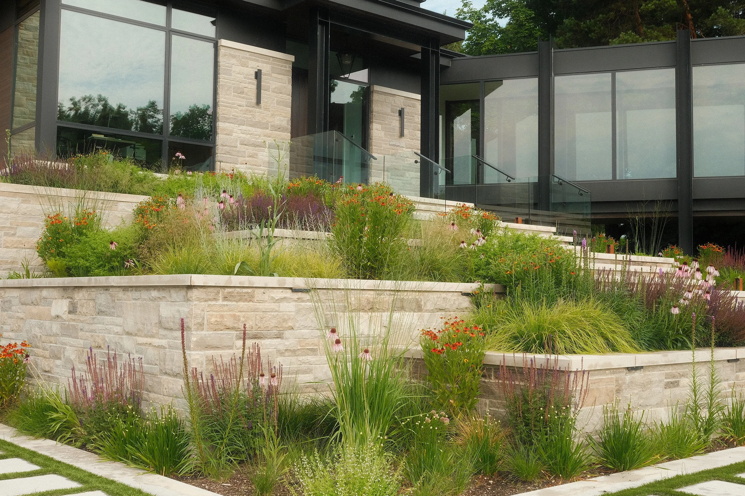 A modern multi-level house with large glass windows and a landscaped terraced garden with various plants and flowers.