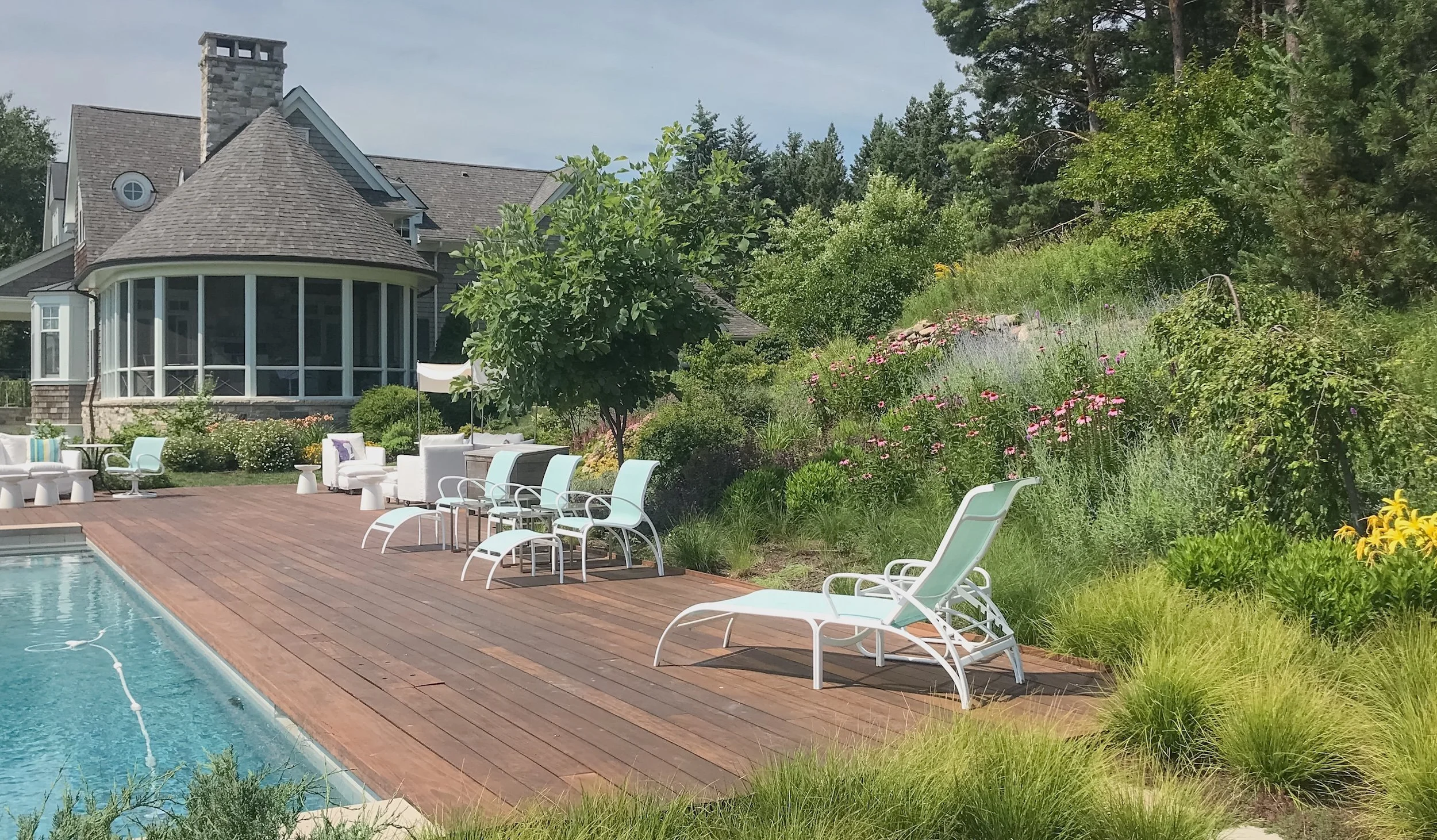 Backyard with a swimming pool, wooden deck, white lounge chairs, a house with a porch, and surrounding lush greenery and flowers.