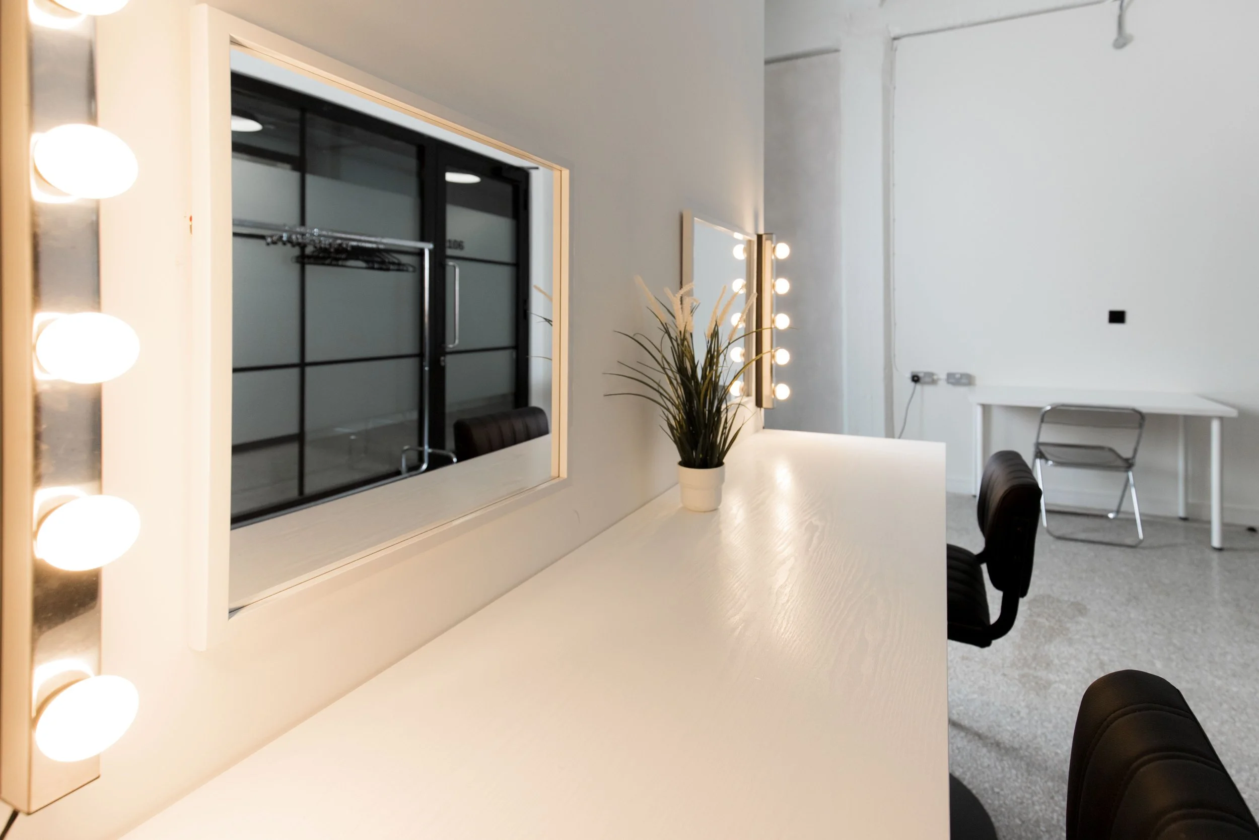 A white dressing table with a mirror bordered by round light bulbs, a potted plant, and black chairs in a room with white walls and a gray floor.