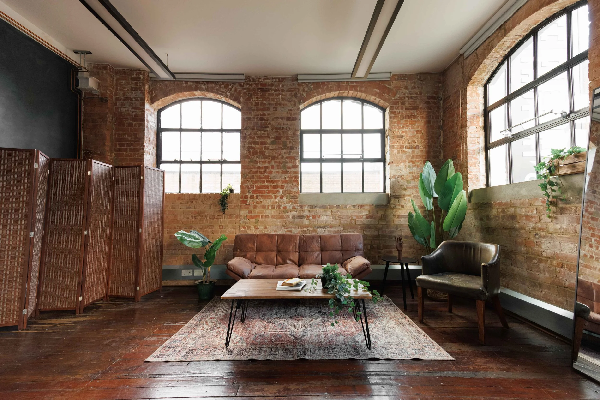 An industrial loft style photography studio with exposed brick walls, large arched windows, a brown leather sofa, vintage armchair, a wooden coffee table, plants, and a make up area on wooden floors. Wood Green, photo and video studio, north london
