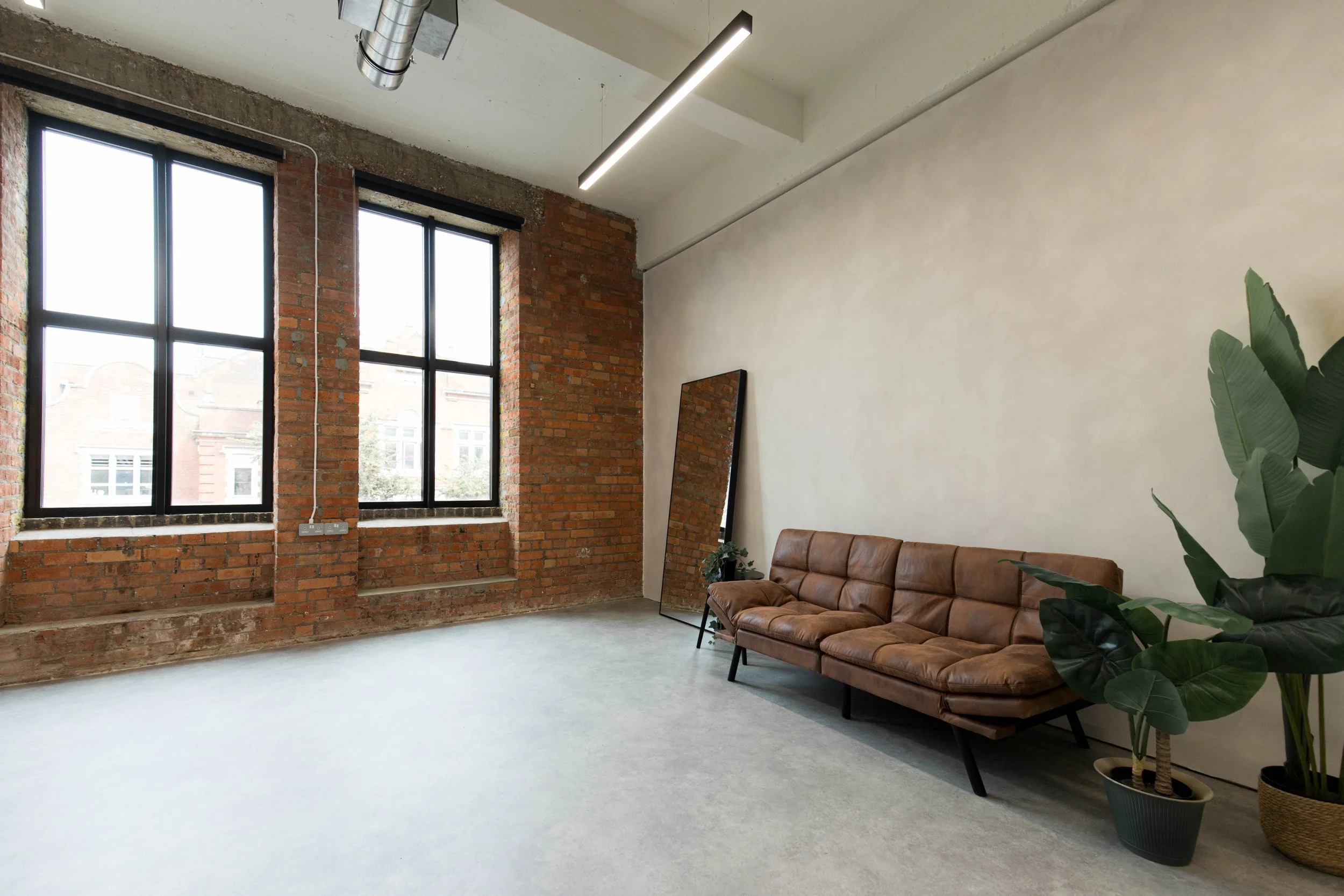 Interior of a modern room with large windows, exposed brick walls, a large mirror resting on the floor, a brown leather couch, and potted plants.
