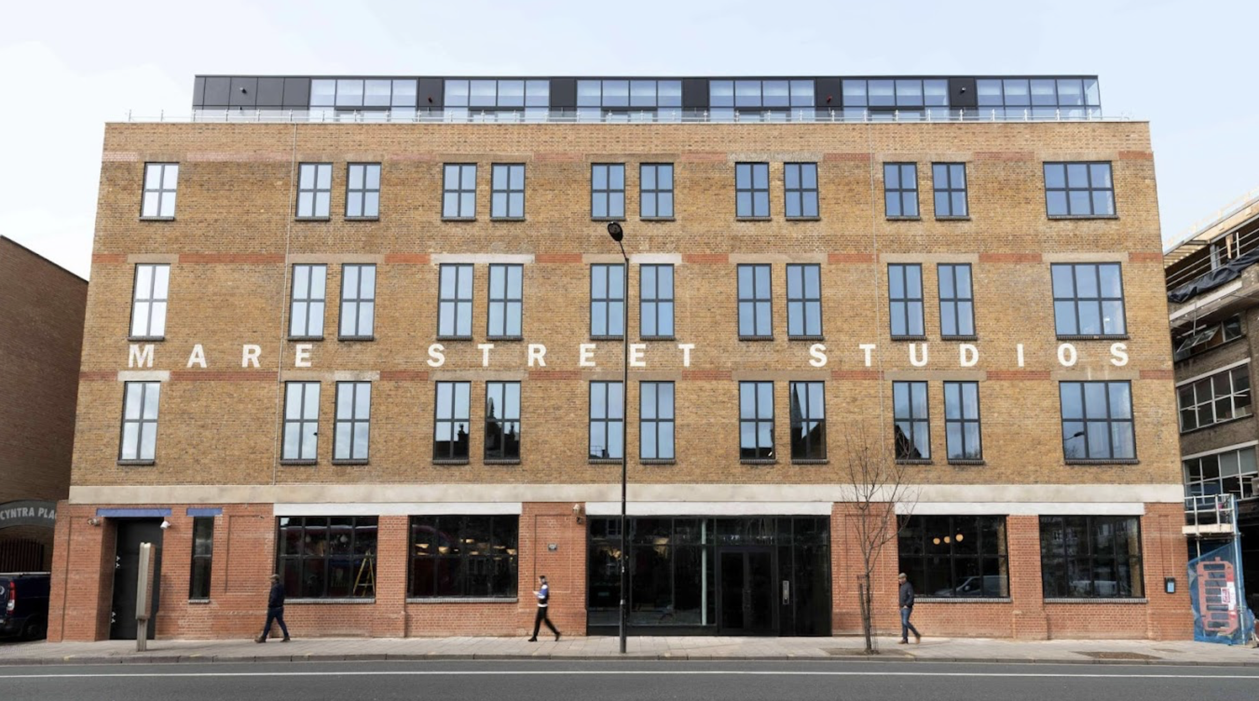 A brick building with large windows and white letters spelling 'MARE STREET STUDIOS' across the facade. There are pedestrians walking on the sidewalk in front of the building, and a streetlamp is visible.