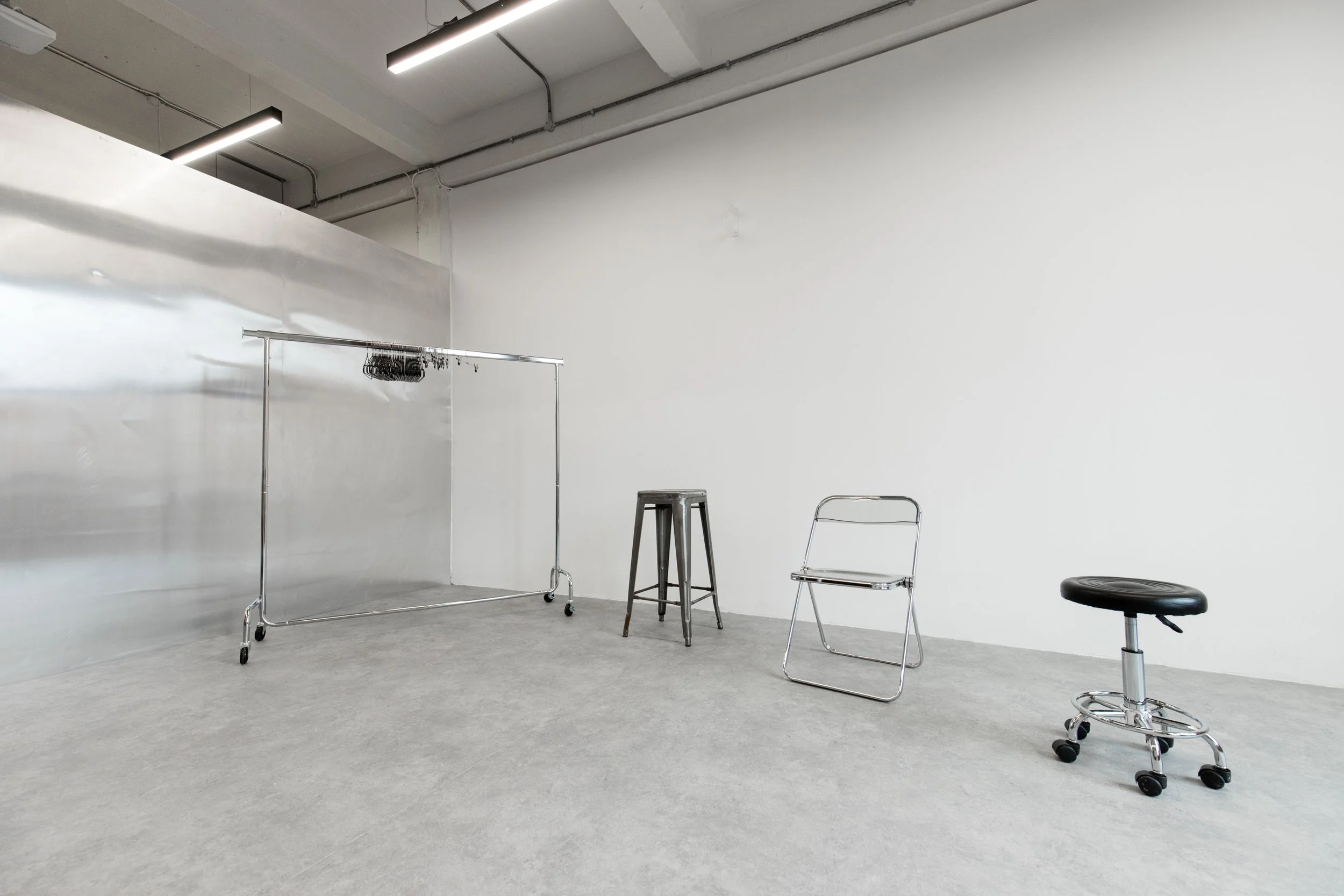 A mostly empty room with a white wall and concrete floor, containing a metal clothing rack, a stool, a chair, and a stool with wheels, metal wall, stainless steel wall, aluminum walls.
