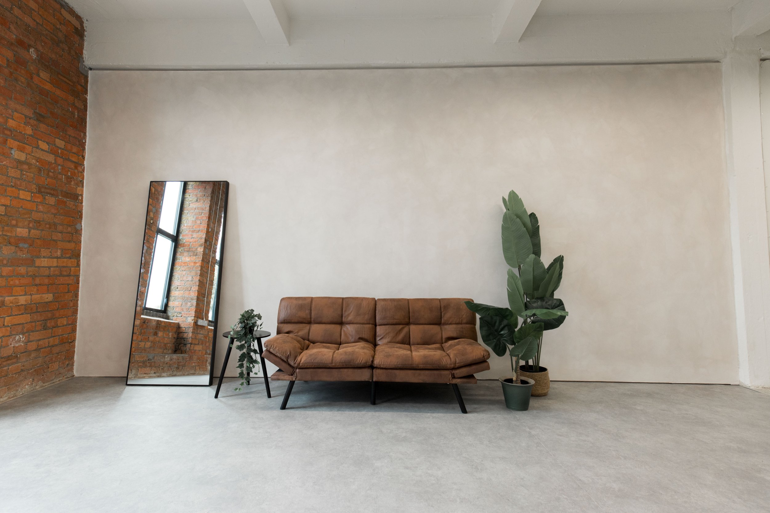 Interior of a room with a brown leather sofa, a tall potted plant, a black side table with decorative items, a large mirror leaning against a white wall, a brick wall on the left, and a window reflected in the mirror.