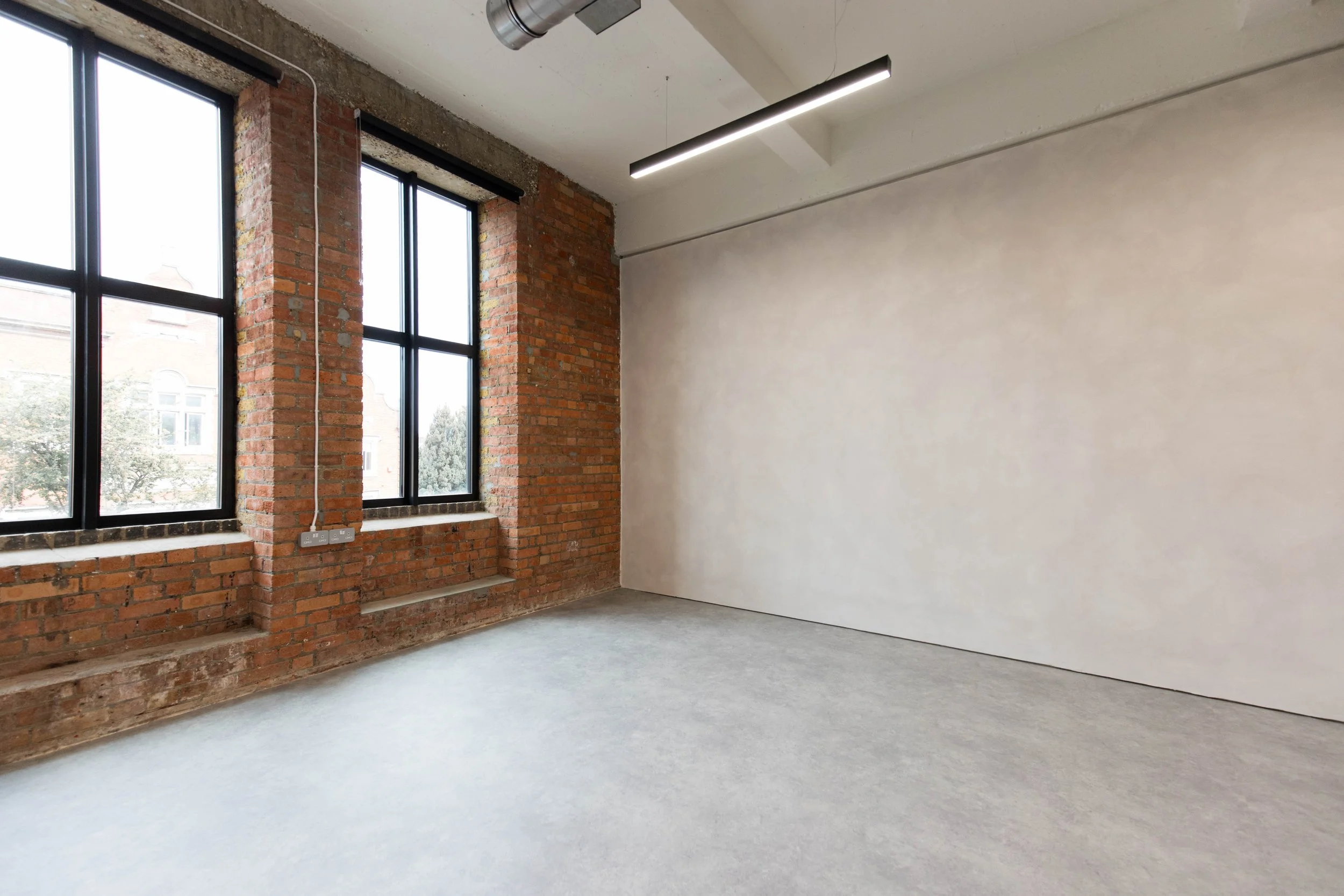 An empty room with large windows framed in black, exposed brick walls, a partial white wall, and a concrete floor. Modern black lighting fixture hanging from the ceiling.