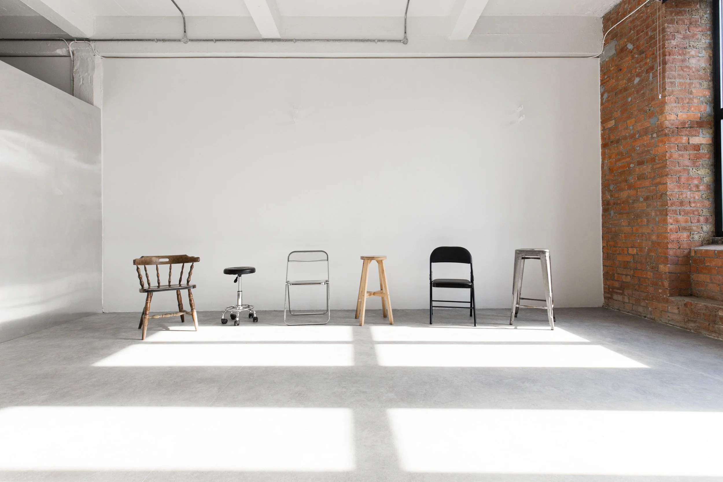 Six chairs of different styles arranged against a white wall in an industrial-style room with exposed brick and pipes.