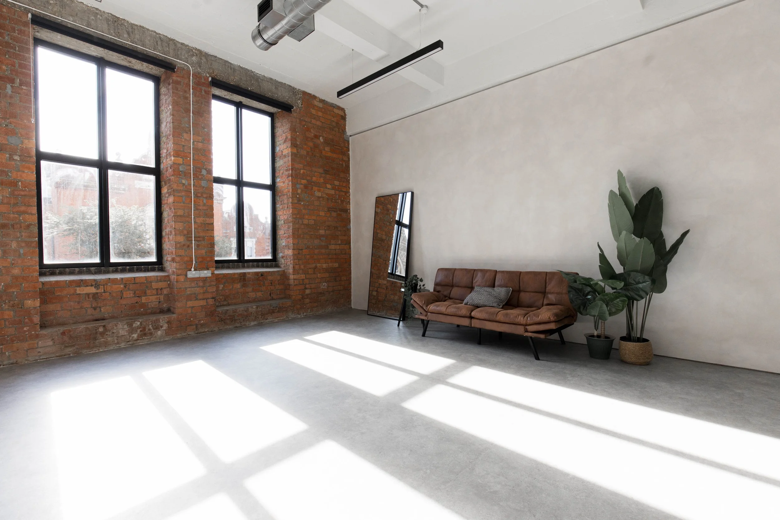 Bright loft-style room with large windows, exposed brick walls, a leather sofa, a tall mirror, and potted plants, with sunlight casting shadows on the concrete floor.