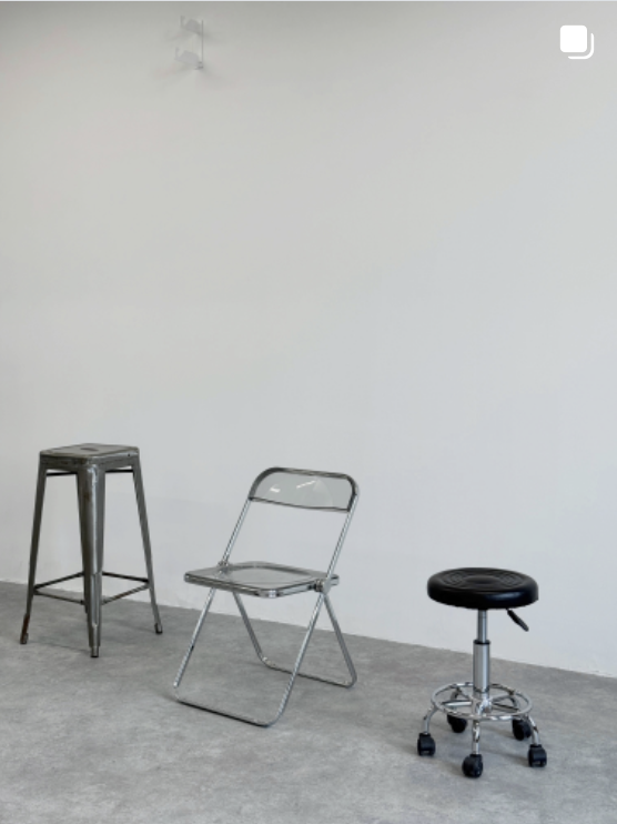 Three different chairs on a grey concrete floor against a plain white wall. One is a tall metal stool, another is a chrome folding chair, and the third is a round black stool with wheels.