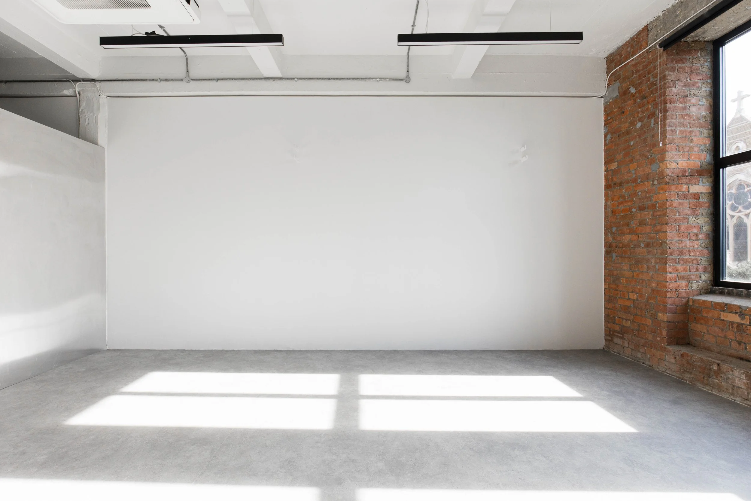 Empty room with white painted walls, natural light from large window with a brick wall, and a concrete floor.