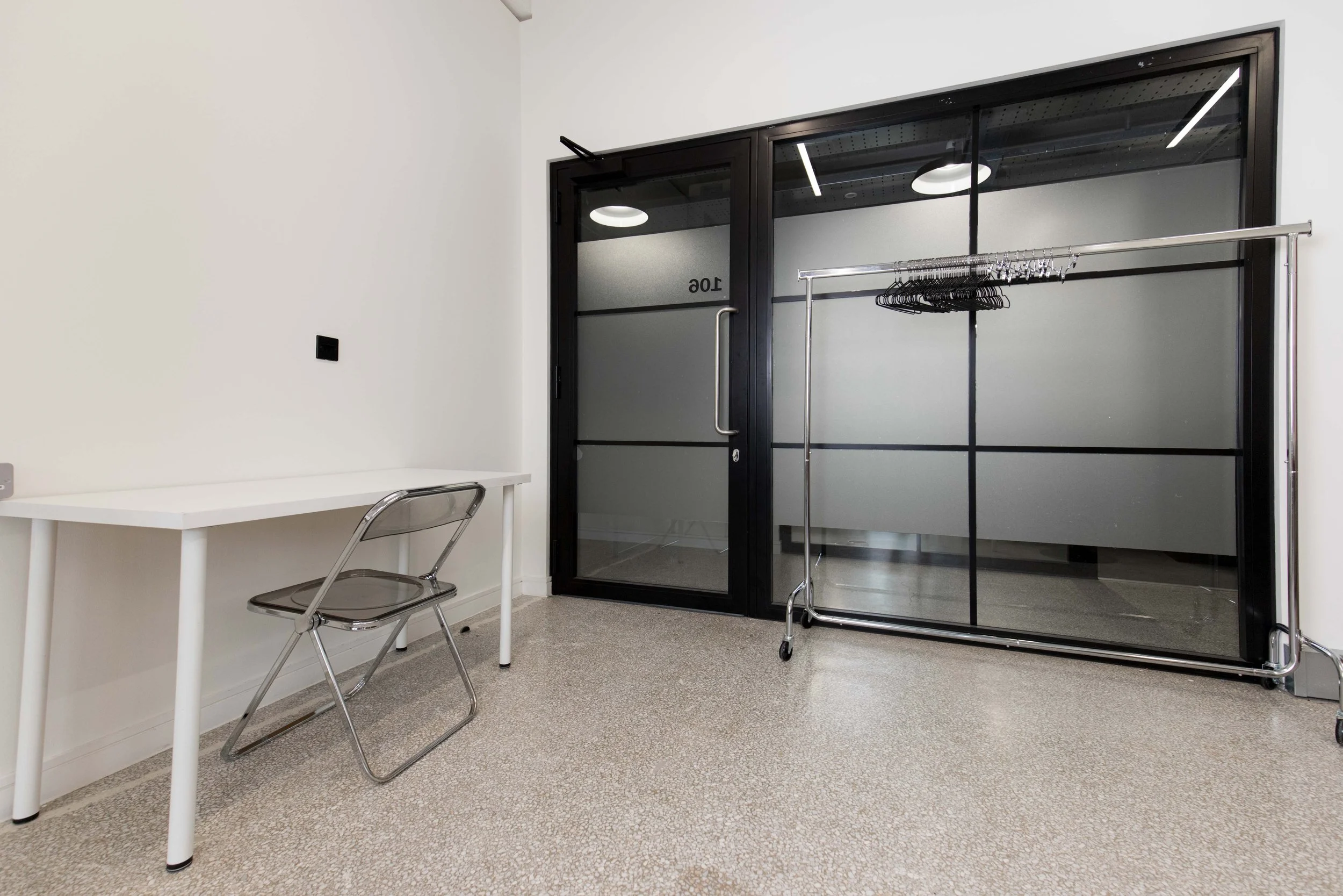 An empty room with a white wall, a white table, a transparent plastic chair, a black framed glass door, and a metal clothing rack with black hangers.