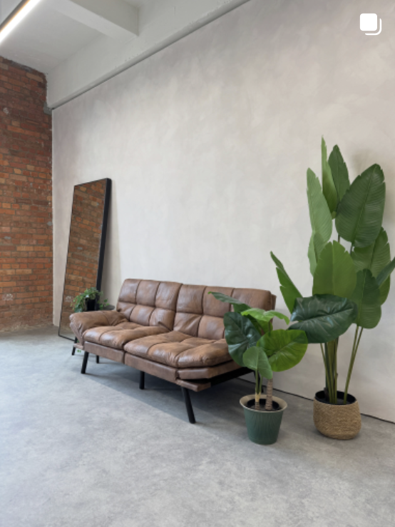 Interior of a minimalist living space with a brown tufted leather sofa, two potted green plants, and a leaning mirror against a white wall, with an exposed brick wall on the left.