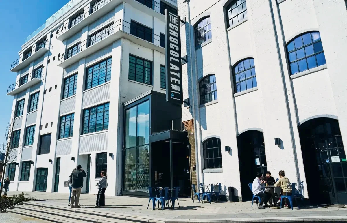 Exterior of a modern multi-story building with a sign reading 'The Chocolate Factory'. Several people are standing and sitting outside, with some engaged in conversation, in front of the building. Wood Green, photo and video studio, north london