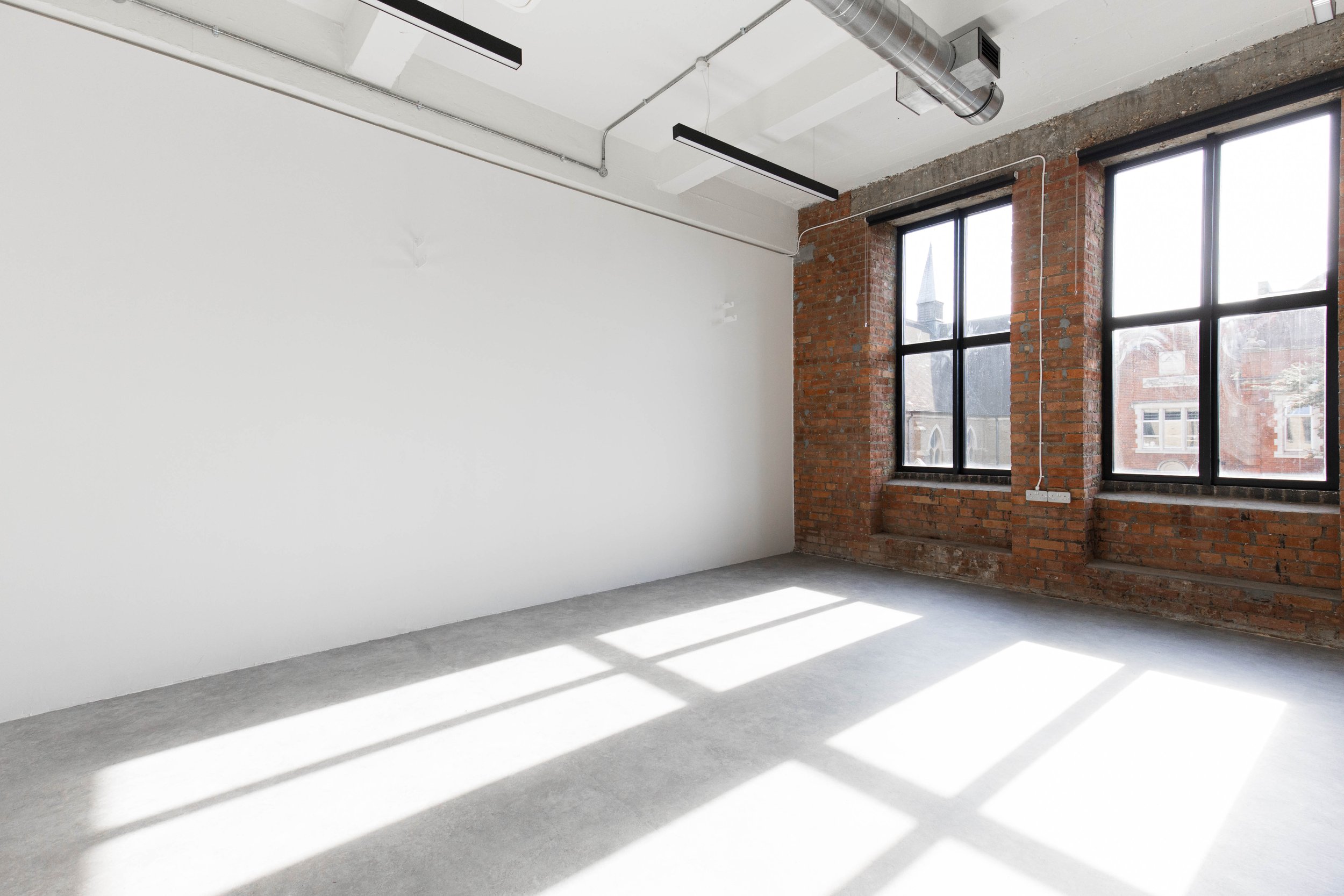 Empty room with white walls, large windows, exposed brick wall, concrete floor, and sunlight creating shadows.
