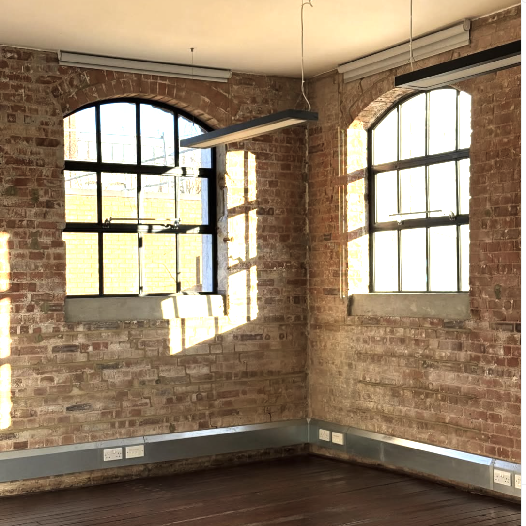 An industrial loft style photography studio with exposed brick walls, large arched windows, a brown leather sofa, vintage armchair, a wooden coffee table, plants, and a make up area on wooden floors. Wood Green, photo and video studio, north london